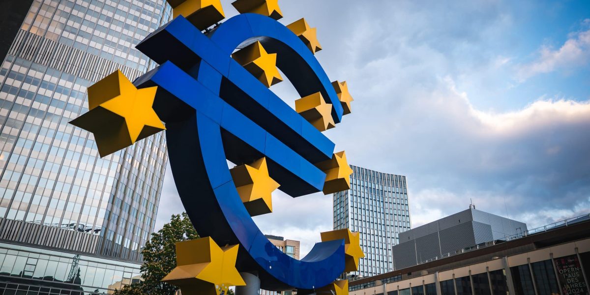 The Euro sculpture in Frankfurt, Germany, surrounded by yellow stars, with modern skyscrapers and the European Central Bank's former headquarters in view.