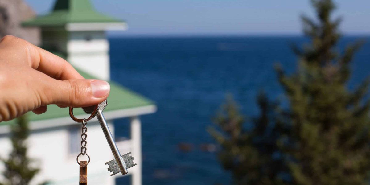 Wooden pendant of a house and key. Background of sea, fir tree and cottage. House and life on ocean, construction, project, moving to new house, mortgage, rent and purchase of real estate. Copy space