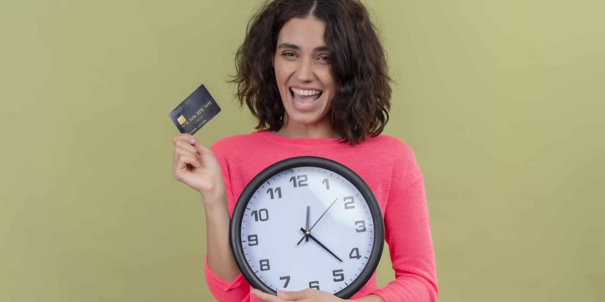 joyful young beautiful girl holding card and clock on isolated green background